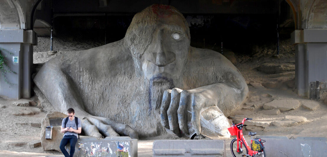 Skulptur eines überdimensionierten Trolls unter einer Brücke.