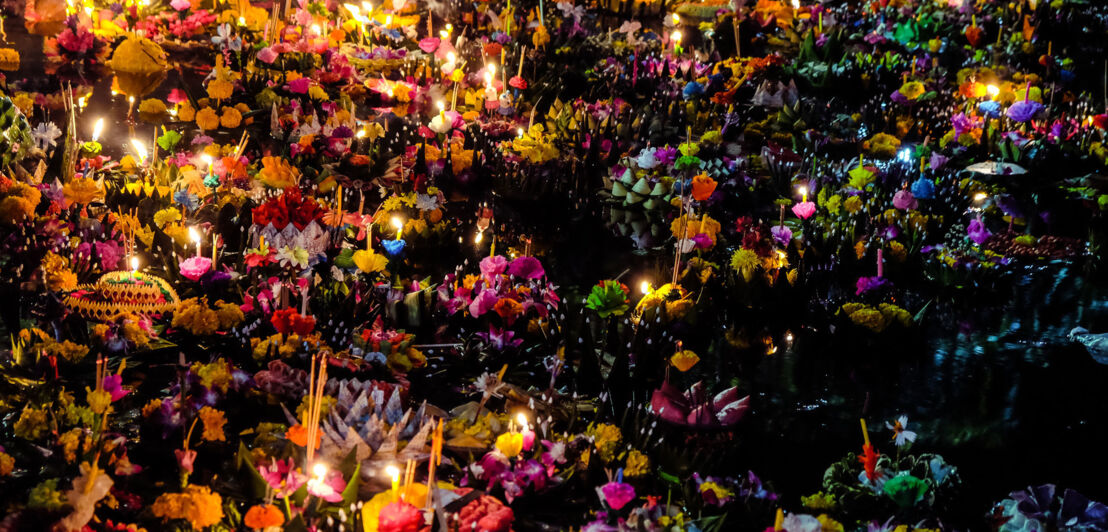 Kleine Flöße üppig geschmückt mit Blumen, Kerzen und Räucherstäbchen im Wasser im Dunkeln.