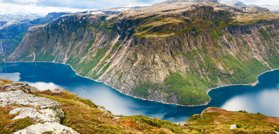 Eisblaues Wasser eines Fjords in Norwegen mit Klippe im Vordergrund und Bergen im Hintergrund.