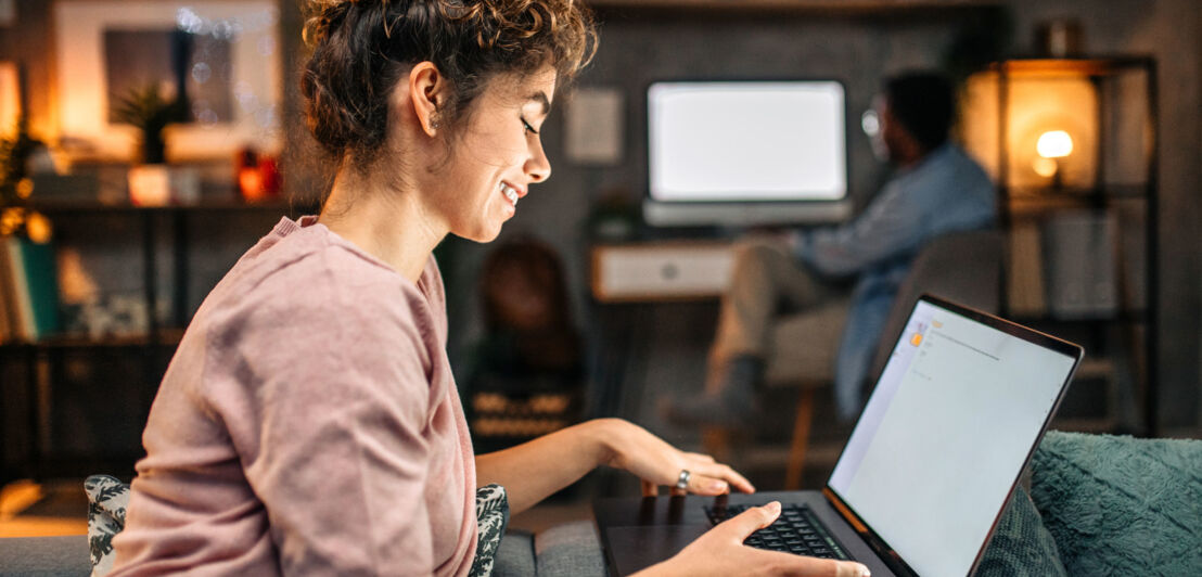 Eine Frau sitzt mit aufgeklapptem Laptop auf einer Couch, im Hintergrund sitzt ein Mann an einem Schreibtisch mit PC.