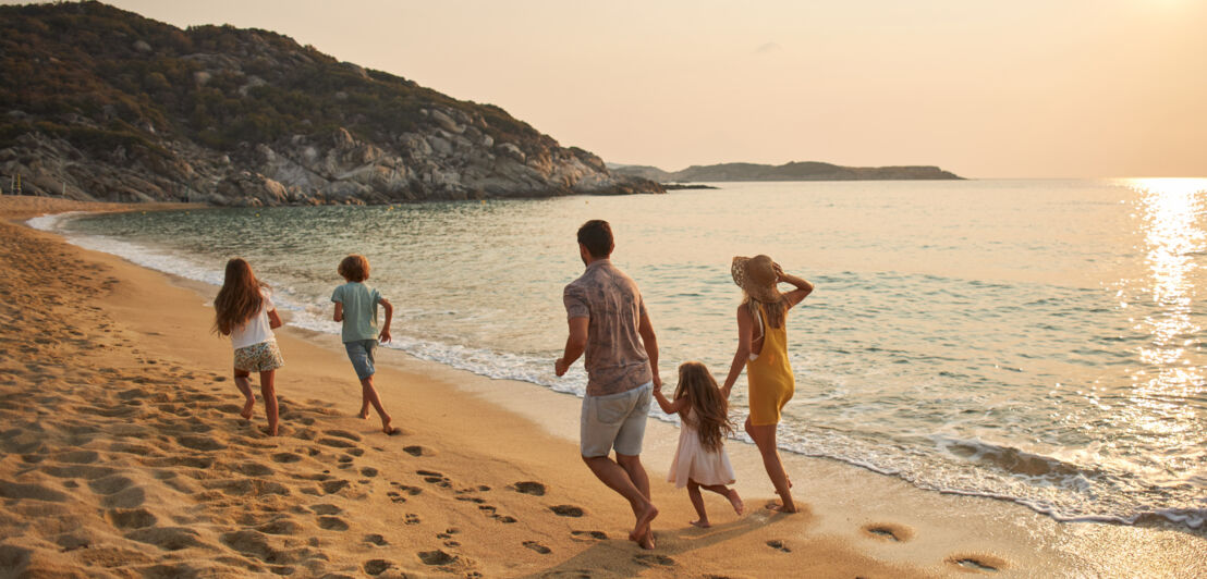 Rückansicht einer Familie mit drei Kindern, die einen Sandstrand entlangläuft.