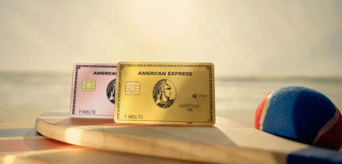 Zwei Kreditkarten von American Express liegen auf einem Holzschläger am Strand, im Hintergrund sieht man das Meer bei Sonnenuntergang. Zwei Kreditkarten von American Express liegen auf einem Holzschläger am Strand, im Hintergrund sieht man das Meer bei Sonnenuntergang.