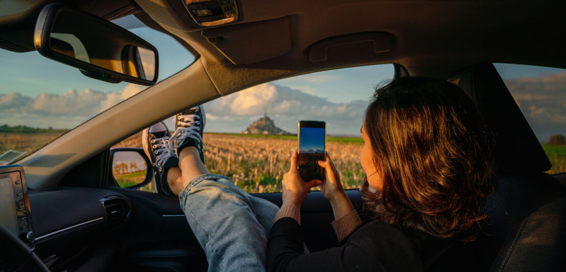 Eine Frau auf dem Beifahrersitz eines Autos hält die Beine aus dem Fenster und fotografiert die Landschaft mit dem Smartphone.