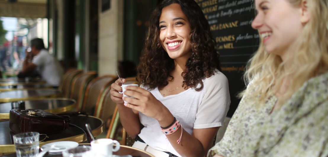 Zwei gut gelaunte Frauen sitzen beim Kaffeetrinken auf der Terrasse eines französischen Bistrots.