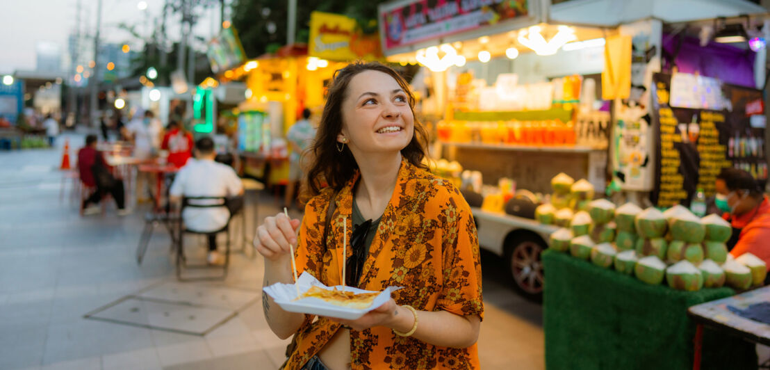 Eine junge Frau mit einer Speise in ihrer Hand vor einem asiatischen Streetfood-Stand.