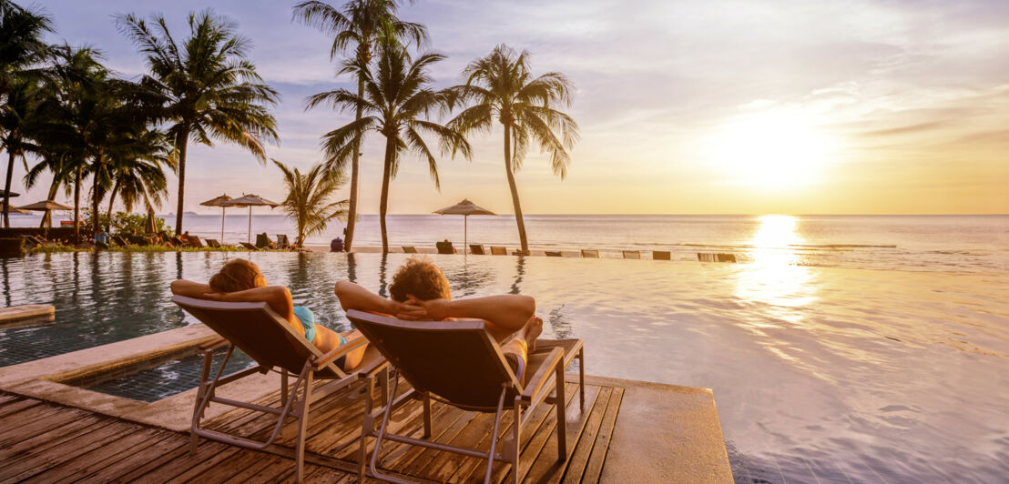 Rückansicht eines Paares in Liegestühlen an einem Hotelpool mit Palmen am Meer bei Sonnenuntergang.