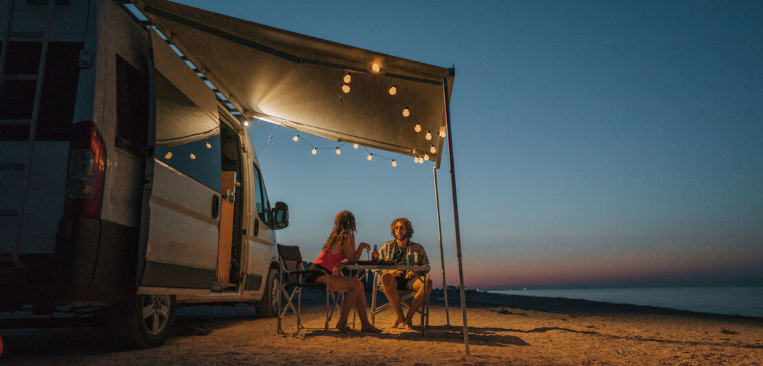 Ein Paar sitzt an einem Campingtisch unter der beleuchteten Markise eines Wohnmobils an einem Sandstrand bei Dunkelheit. Ein Paar sitzt an einem Campingtisch unter der beleuchteten Markise eines Wohnmobils an einem Sandstrand bei Dunkelheit.