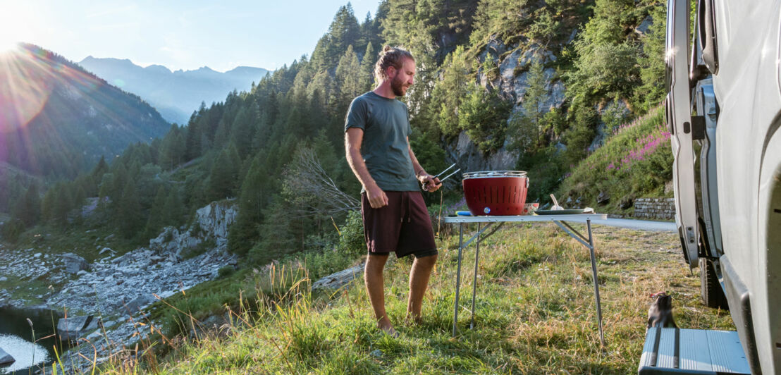 Ein Mann steht mit einer Grillzange an einem Tisch mit tragbarem Grill neben einem Wohnmobil in einer Berglandschaft. Ein Mann steht mit einer Grillzange an einem Tisch mit tragbarem Grill neben einem Wohnmobil in einer Berglandschaft.