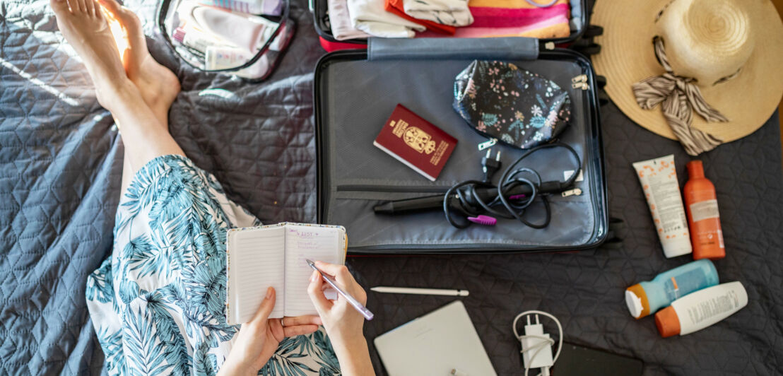 Aufsicht eines Koffers und ausgebreiteten Reiseutensilien auf einem Bett, daneben Beine und Hände einer Frau, die in ein Notizheft schreibt. Aufsicht eines Koffers und ausgebreiteten Reiseutensilien auf einem Bett, daneben Beine und Hände einer Frau, die in ein Notizheft schreibt.