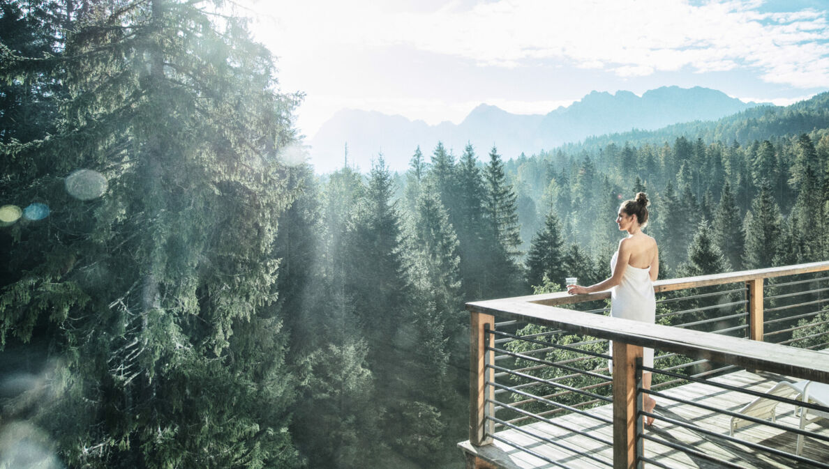 Eine in ein Handtuch gewickelte Frau blickt von einer Terrasse auf einen herrlichen Nadelwald