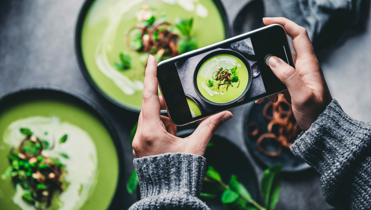 Zwei Hände halten Smartphone, das eine grüne Suppe in Schüssel fotografiert