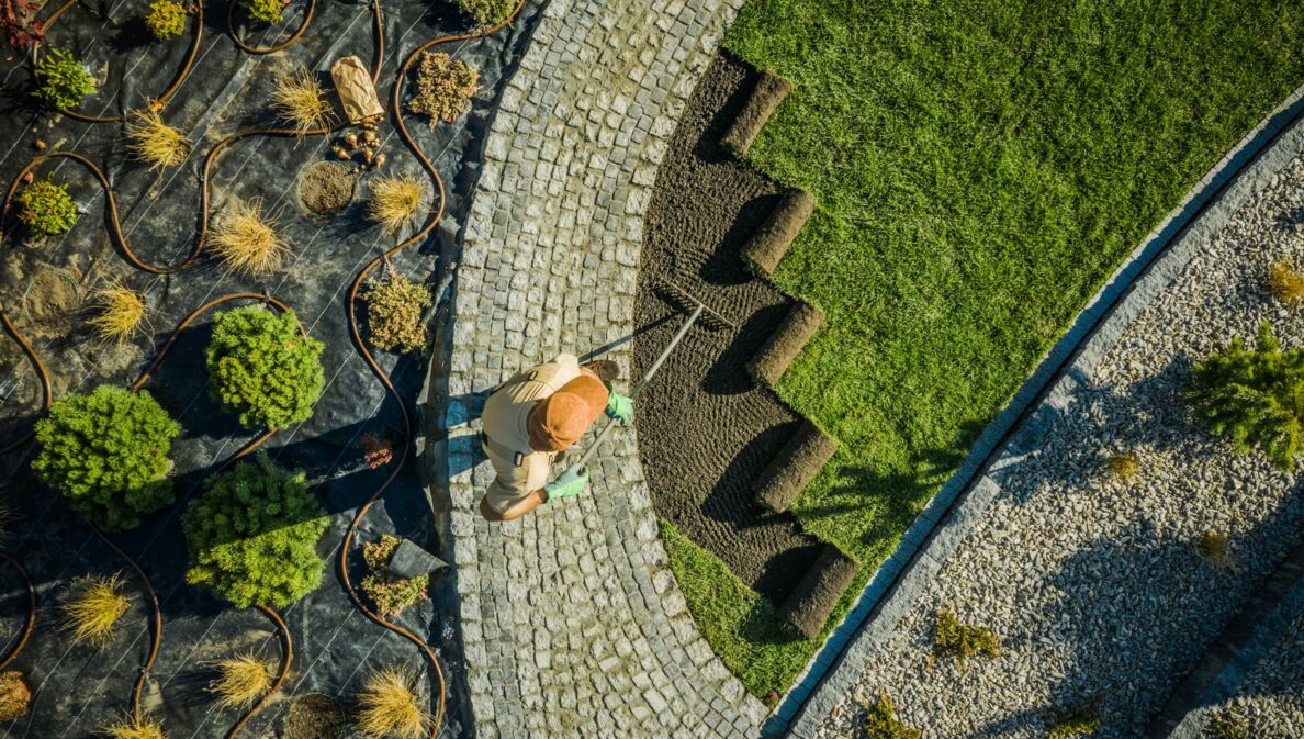 Luftaufnahme einer Person, die auf einem gepflasterten Weg stehend eine Fläche mit Rollrasen in einem Garten bearbeitet