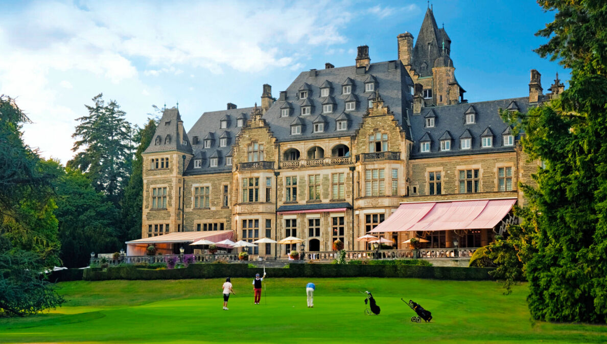 Außenaufnahme von Schlosshotel mit Terrasse, davor Rasen mit Golfenden