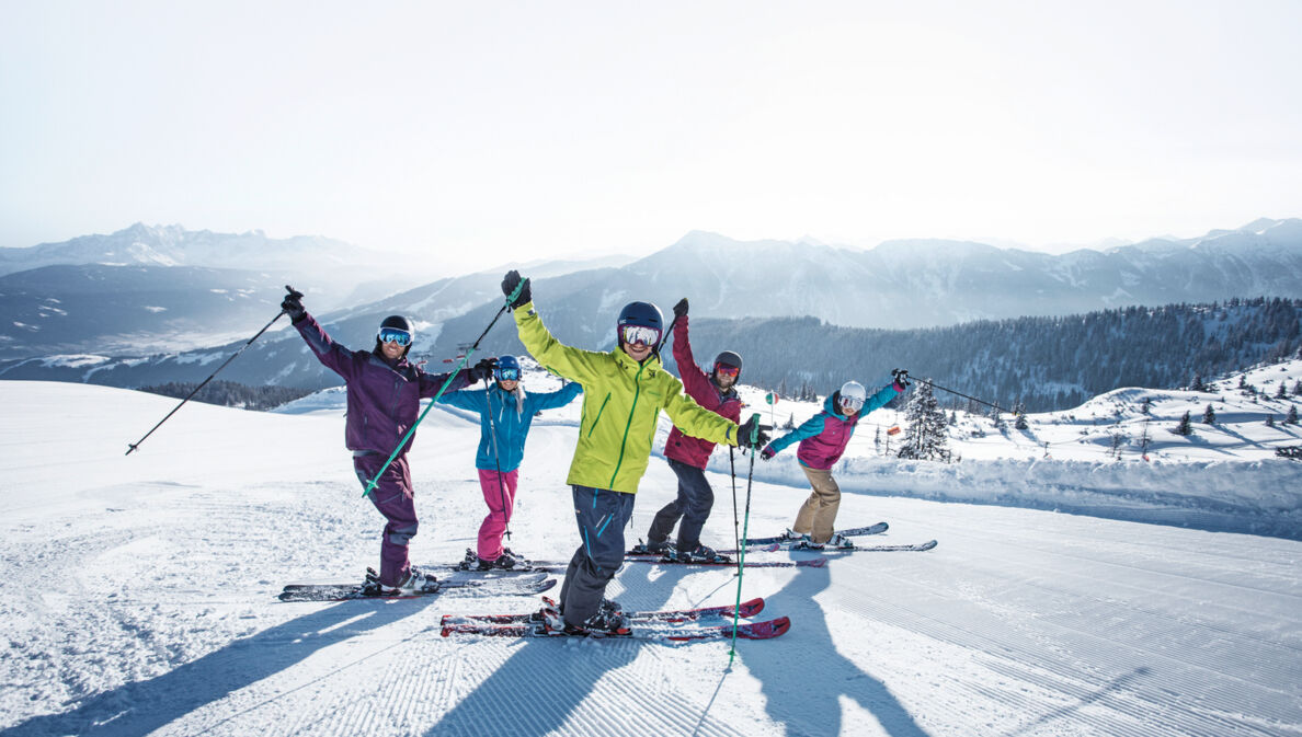 Fünf Skifahrende in bunter Kleidung winken in die Kamera auf einer Piste vor winterlichem Bergpanorama