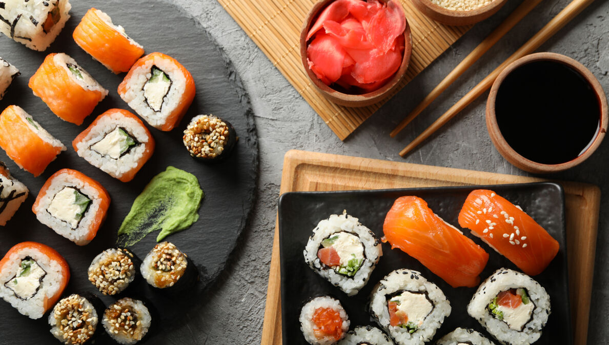 Aufsicht unterschiedlicher Sushi-Sorten auf zwei Tellern, daneben Beilagen und Stäbchen