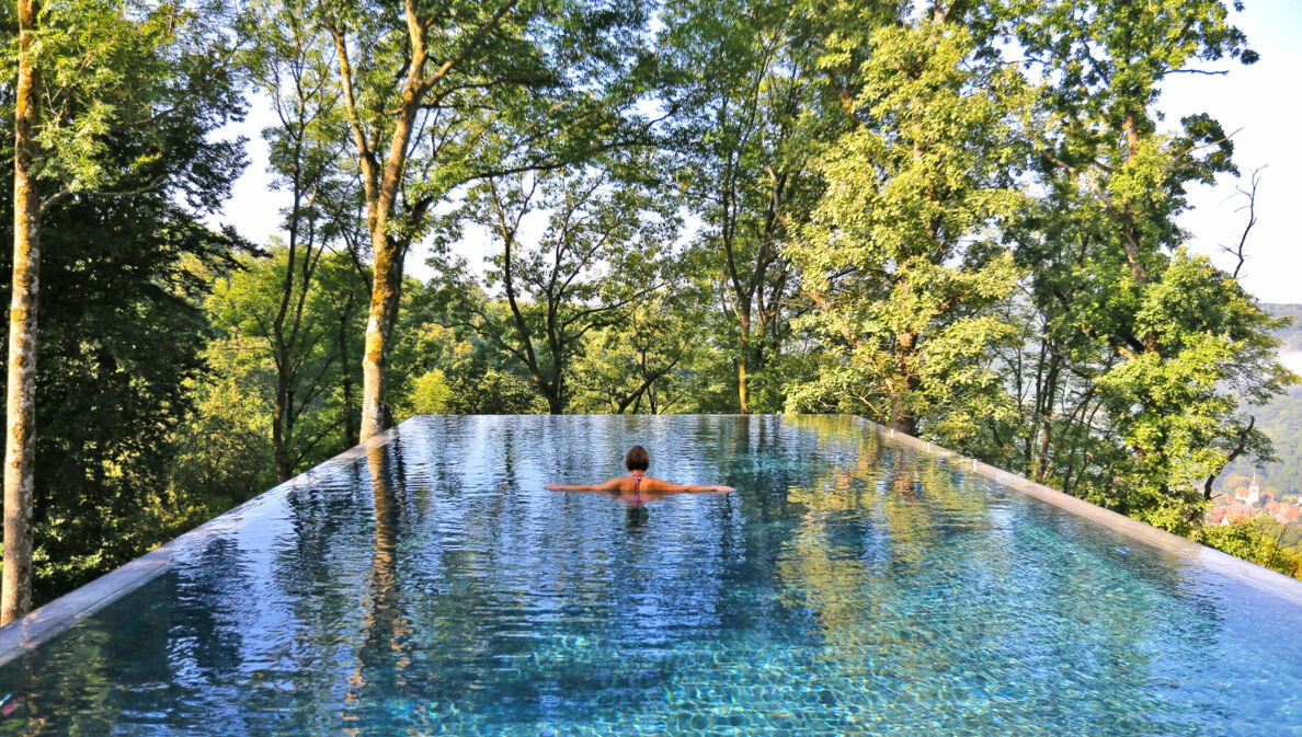 Rückansicht einer Frau im Infinity-Pool, im Hintergrund Baumkronen