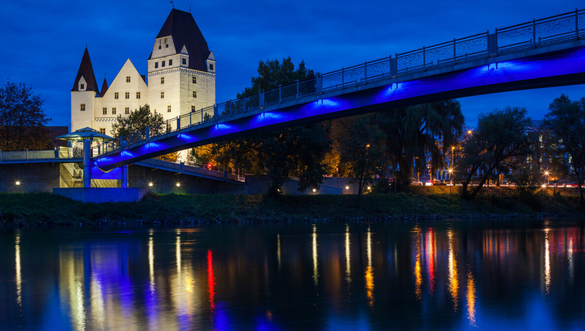 Das beleuchtete Neue Schloss in Ingolstadt bei Nacht, im Vordergrund führt eine Brücke über die Donau