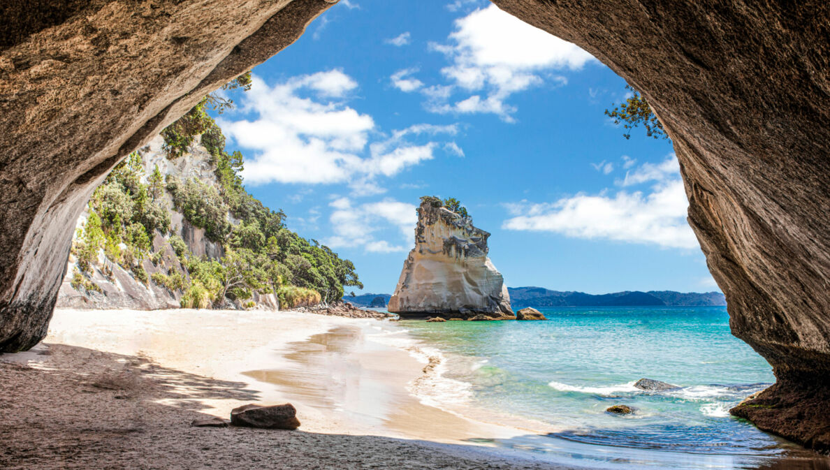 Im Vordergrund ein Felsbrocken, dahinter ein Sandstrand, umringt von Felsen und türkisfarbenem Meer