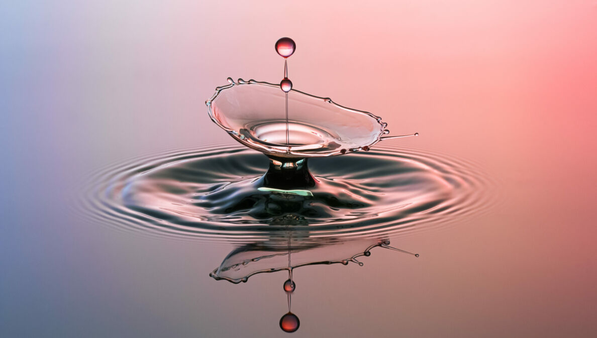 Eine lilafarben und rot angestrahlte Wasserfläche, in die ein Wassertropfen fällt.