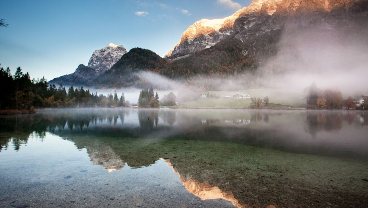 Ein See mit glasklarem Wasser über dem Nebelschwaden liegen vor Bergpanorama in der Dämmerung