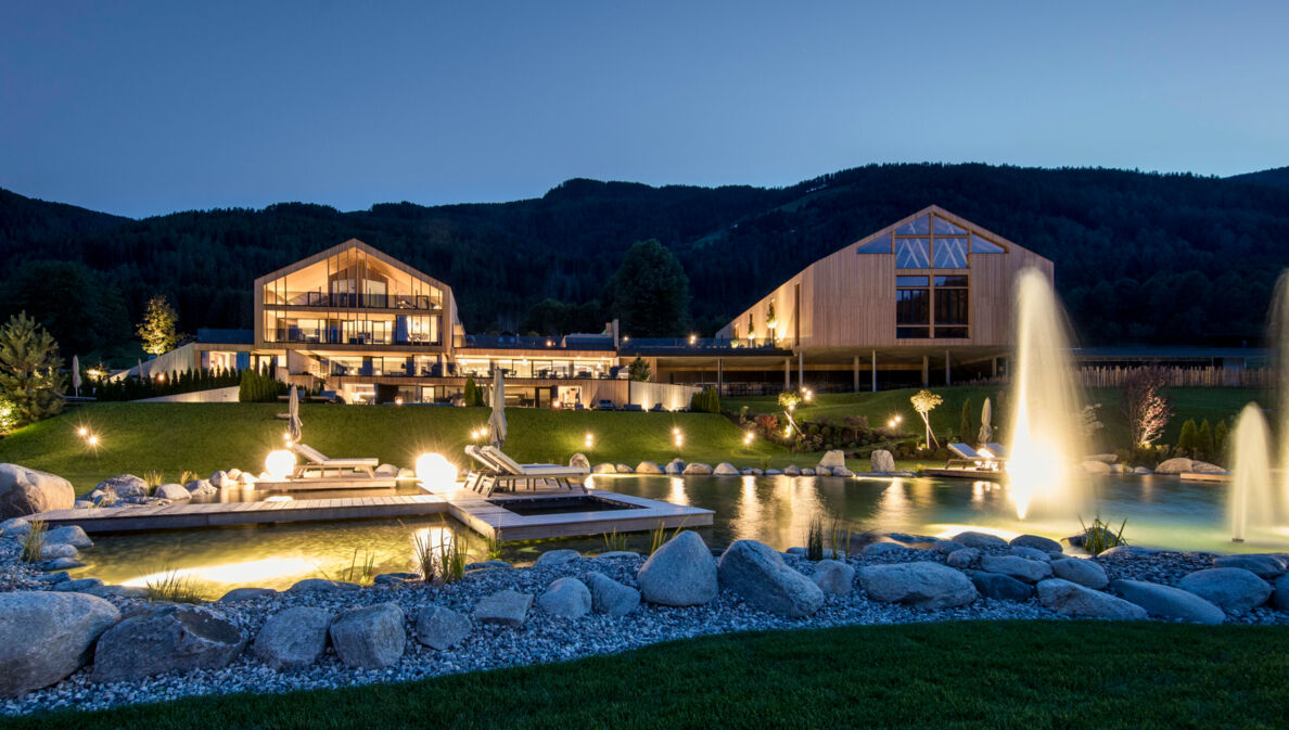 Eine luxuriöse, beleuchtete Hotelanlage aus Holzgebäuden mit Landschaftsgarten und Badeteich vor Bergpanorama bei Nacht