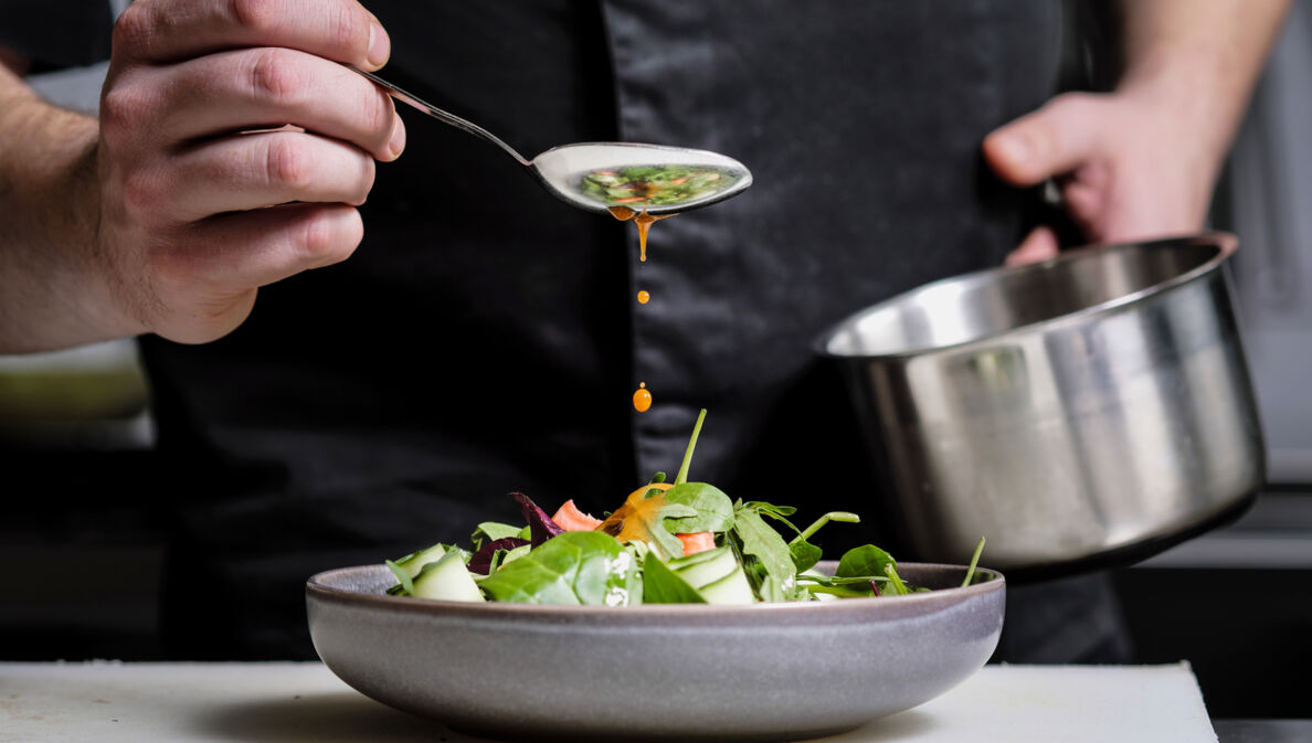 Nahaufnahme einer Hand, die mit einem Löffel gelbes Dressing über einen Teller mit Rohkost träufelt