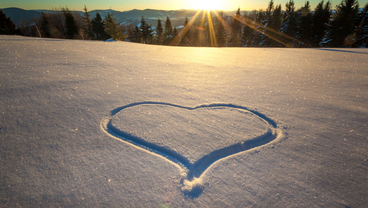 Ein Herz in den Schnee gezeichnet, im Hintergrund ein Wald und Berge