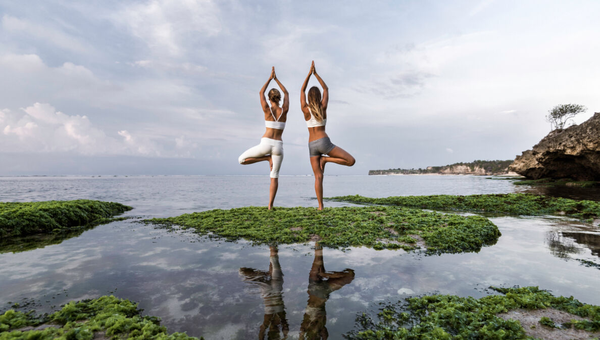 Zwei Frauen stehen am Wasser und machen zwei Yogaübungen