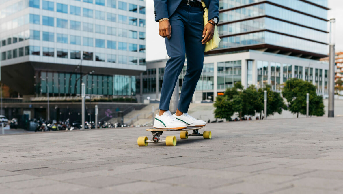 Zwei Beine auf einem Skateboard, die Person trägt eine Anzughose und weiße Sneaker