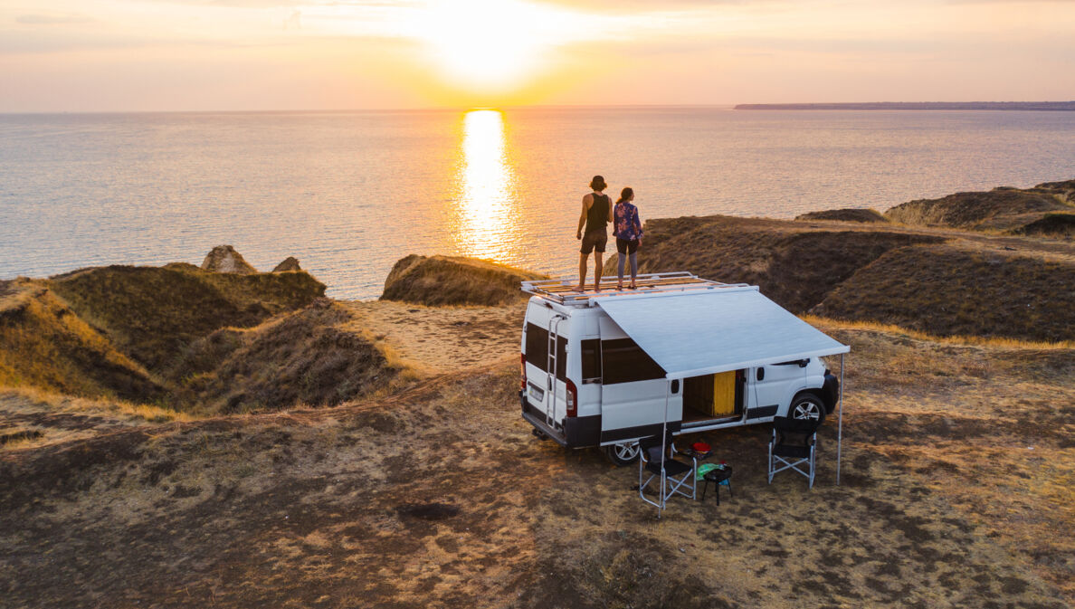 Ein Paar auf dem Dach eines Wohnmobils mit Blick auf die tiefstehende Sonne über dem Meer