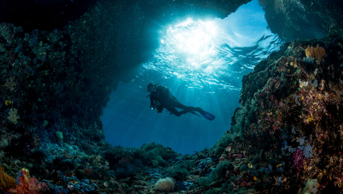 Taucher unter Wasser am Eingang einer Höhle mit Sonnenstrahlen im Hintergrund