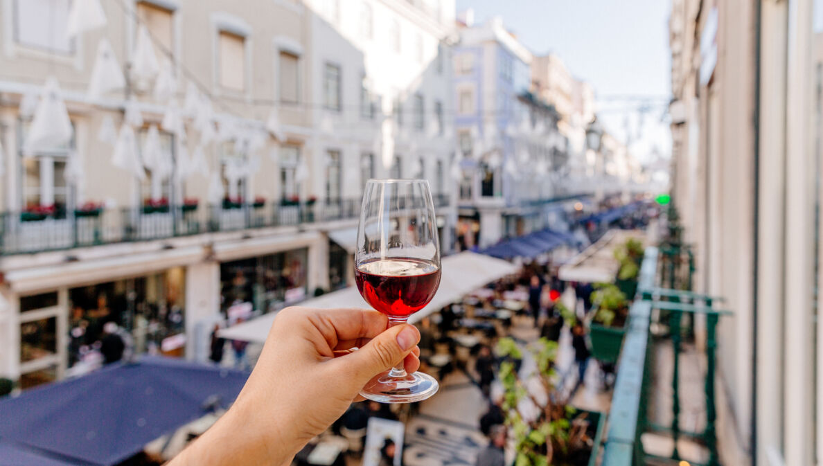 Eine Hand hält ein Glas Portwein oberhalb einer belebten Gasse in die Luft