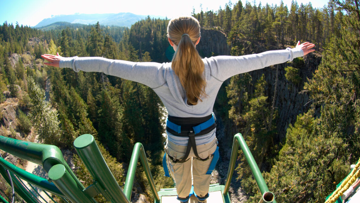 Eine Frau steht mit ausgebreiteten Armen gesichert an einem Bungee-Jumping-Gerüst und ist kurz davor runterzuspringen