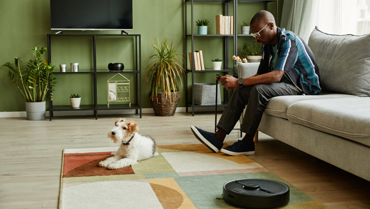 Ein Mann sitzt im Wohnzimmer auf dem Sofa und schaut auf sein Handy in der Hand. Vor ihm liegt ein Hund auf dem Teppich, daneben arbeitet ein Saugroboter.