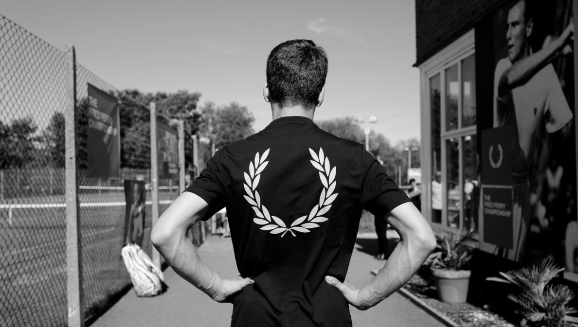 Rückansicht eines Mannes in einem dunklen T-Shirt mit Lorbeerkranz-Logo-Druck auf dem Rücken auf einem Tennisplatz, Schwarzweißaufnahme