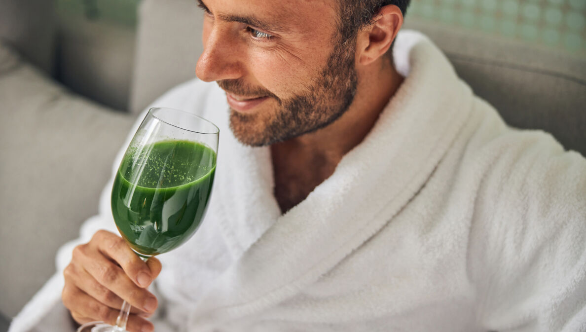 Mann in Bademantel, der ein Glas mit grünem Inhalt in der Hand hält