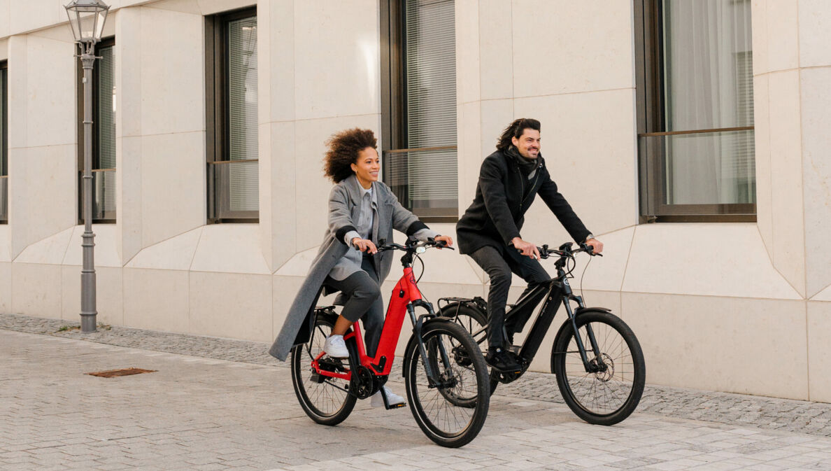 Ein Mann und eine Frau radeln auf zwei E-Bikes an einem Haus vorbei