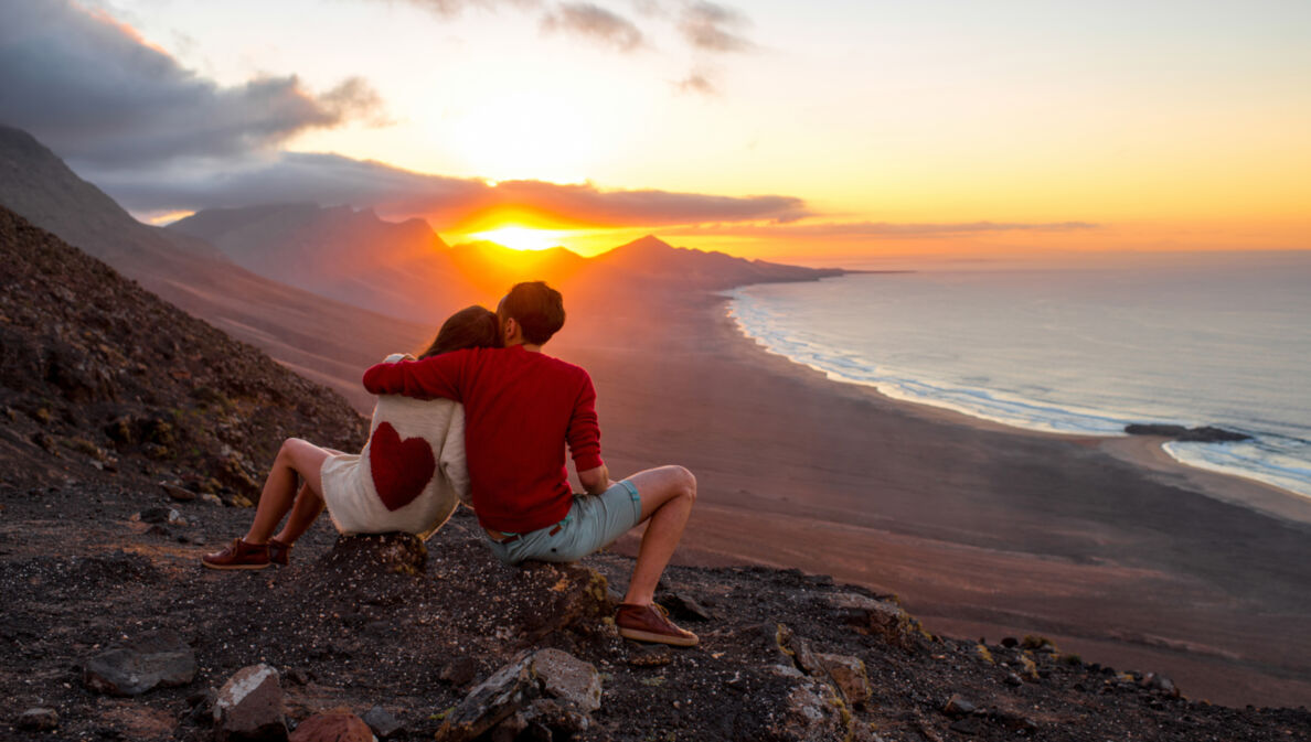 Zwei Personen sitzen mit dem Rücken zur Kamera auf einem Felshang und Blicken in den Sonnenuntergang über den Bergen, rechts im Bild das Meer