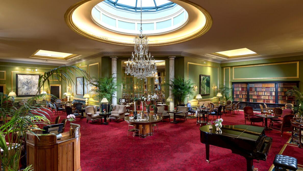 Elegante Hotellobby mit Kronleuchter und rotem Teppichboden, auf dem sich ein Piano und Sitzgelegenheiten befinden