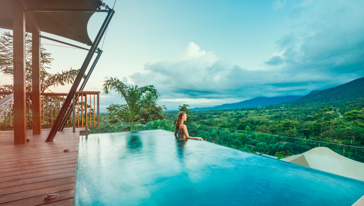 Profilansicht einer Frau, die aus einem Infinity-Pool über eine tropische Hügellandschaft schaut