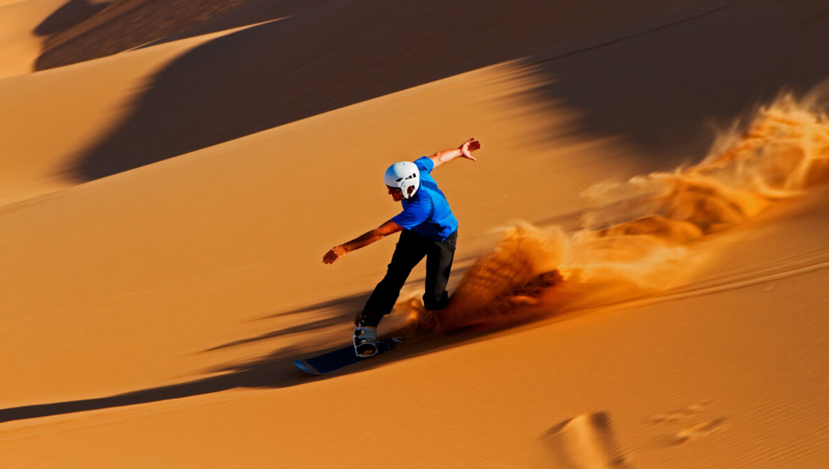 Ein Sandboarder mit Helm fährt eine rötliche Sanddüne hinunter