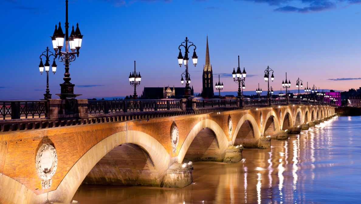 Eine alte Steinbrücke mit erleuchteten Straßenlaternen verläuft über einen Fluss in Bordeaux bei Nacht