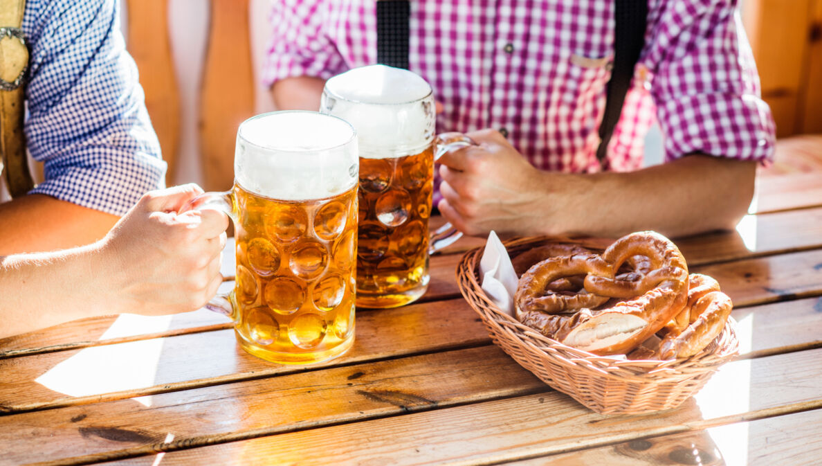 Nahaufnahme von zwei Männern, die an einem Tisch in einem Brauhaus mit zwei Maßkrügen Bier anstoßen