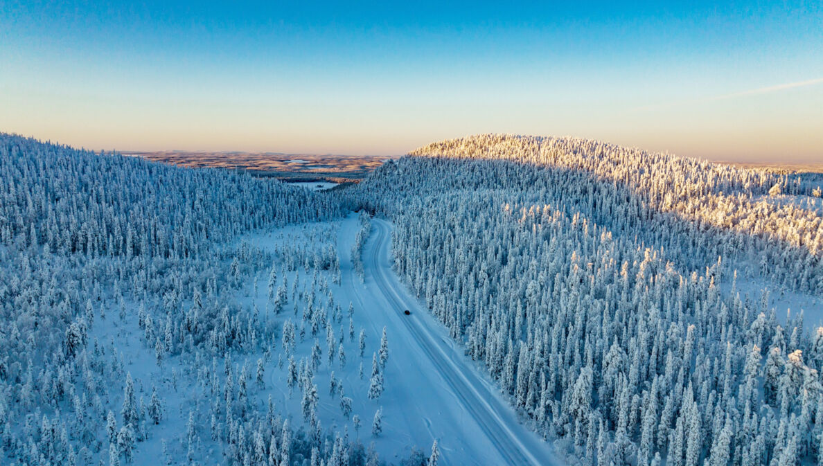 Luftaufnahme einer weiten Winterlandschaft mit schneebedeckten Wäldern, durch die ein einzelnes Auto über eine Straße durch den Schnee in der Abenddämmerung fährt