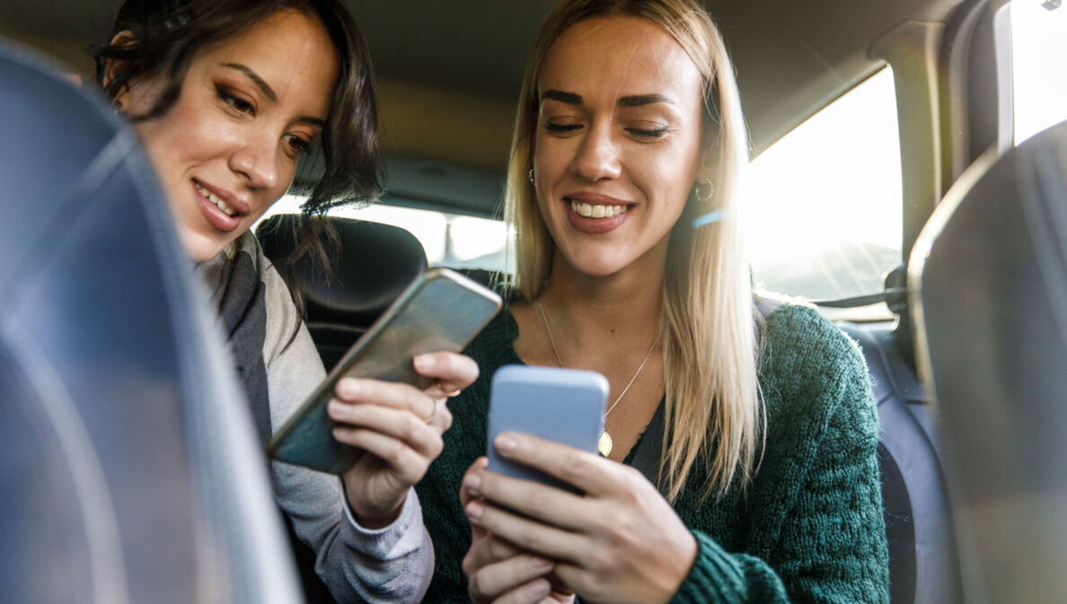 Zwei junge Frauen auf der Rückbank eines Autos schauen lächelnd auf ihre Smartphones