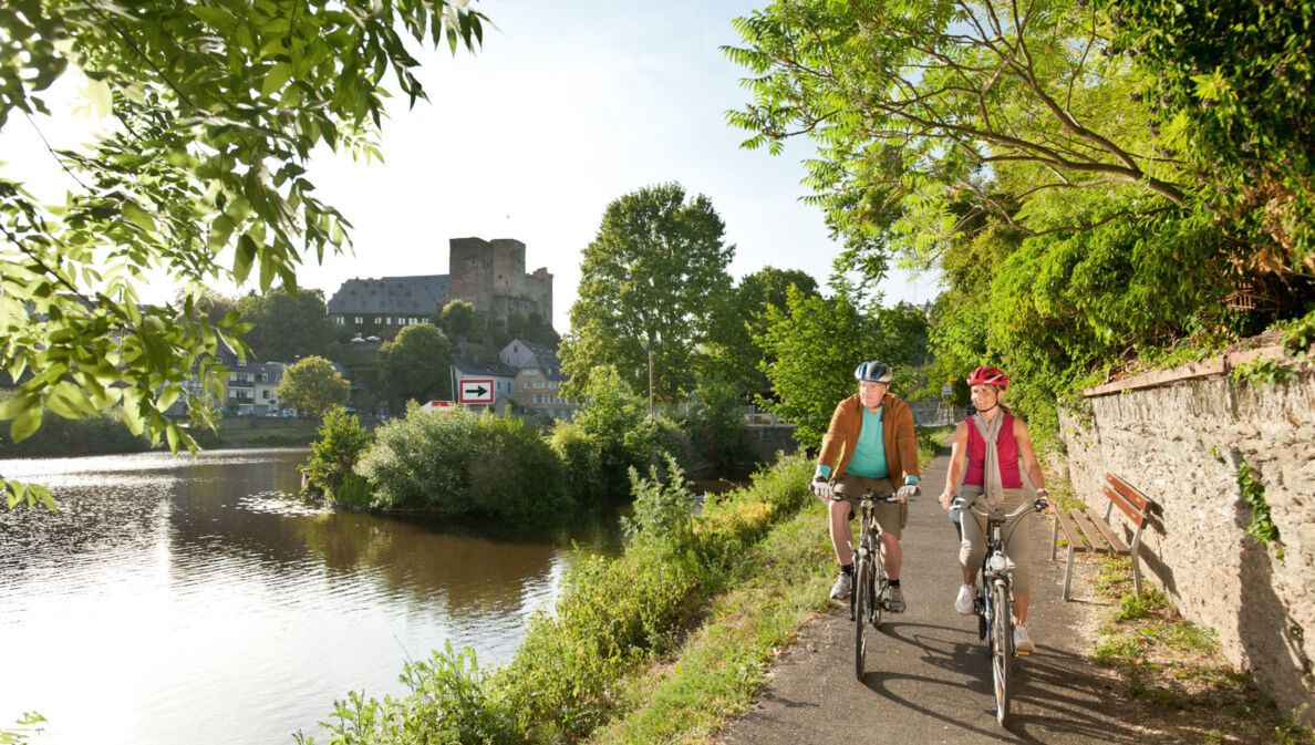 Ein älteres Paar fährt auf Fahrrädern auf einem asphaltierten Radweg entlang eines Flusses, im Hintergrund eine mittelalterliche Burgruine in einer Ortschaft