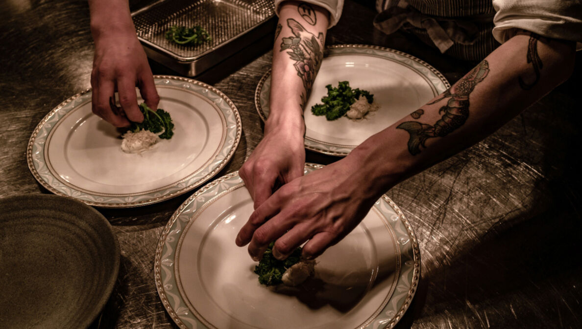 Nahaufnahme von Händen beim Anrichten von Speisen auf großen, weißen Tellern in einer Restaurantküche.