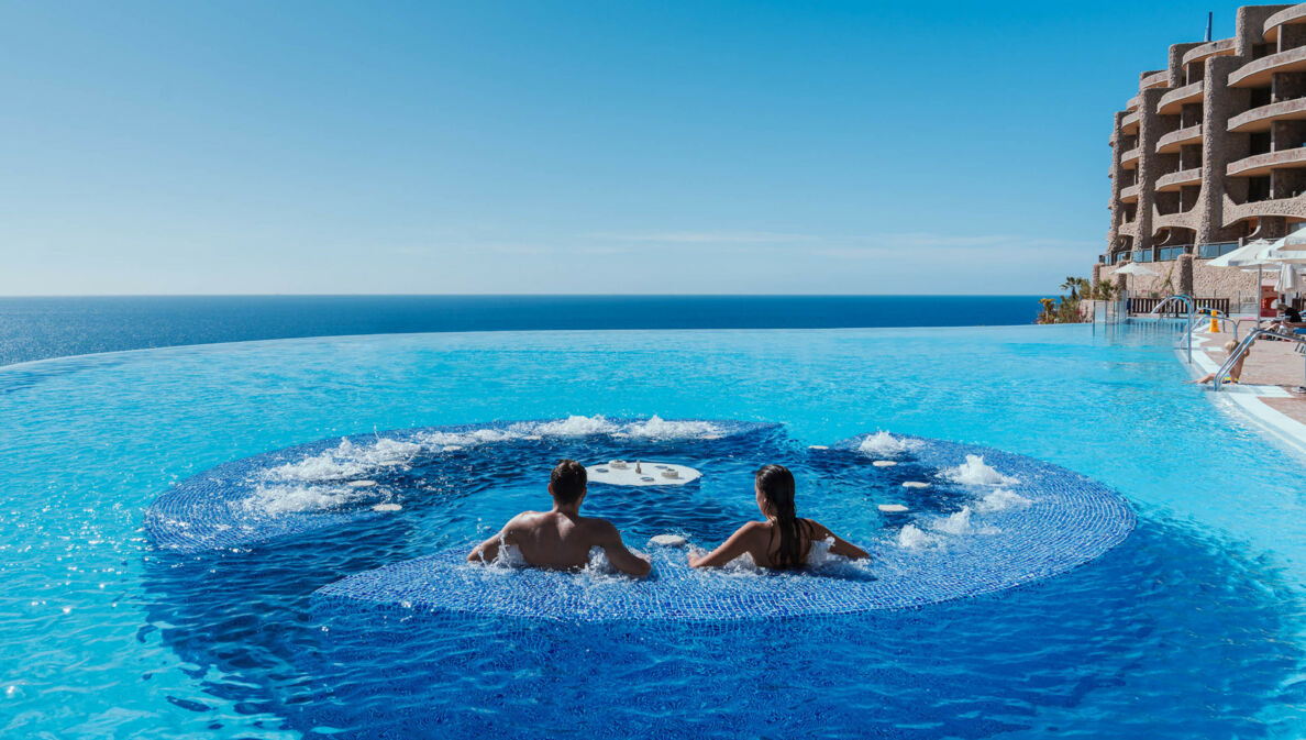 Rückansicht zweier Personen in einem großen Hotelpool mit Jacuzzi am Meer.