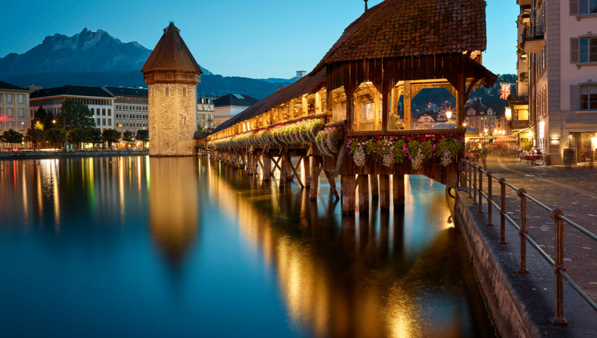 Eine überdachte und beleuchtete Holzbrücke über einem Fluss, ein Wasserturm und eine Stadt und Berge im Hintergrund.