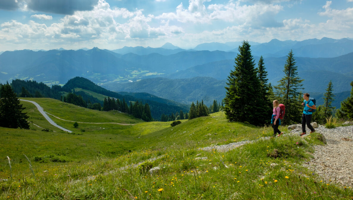 Ein wanderndes Paar mit Rucksäcken auf einem Weg inmitten einer grünen Berglandschaft.